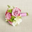A small, hand-tied bouquet lies horizontally on a pale, textured linen surface, its colours glowing softly as if caught in gentle afternoon light over Golders Green. At the centre, two fully open roses in a dusty blush pink unfold layer upon layer of velvety petals, their outer edges deepening to a richer magenta that frames each bloom with a delicate ombré halo. A third rose, slightly smaller and more tightly furled, nestles nearby, suggesting new petals still to open. Around them, round tufts of lime-green button chrysanthemums form playful pom-pom shapes, their tiny packed florets adding a crisp, modern contrast to the romantic roses. Threads of creamy white filler flowers, reminiscent of lisianthus or freesia, weave between the larger heads, their soft, ruffled petals and tiny starry blossoms lightening the composition and filling any gaps with airiness. Slender hints of deep green foliage peek out, giving structure and a subtle woodland freshness. The stems are gathered into a neat posy and wrapped in luminous fuchsia satin ribbon, the handle smooth and slightly reflective, like evening light on shopfronts along Golders Green Road. Over the ribbon, a fine strand of small white faux pearls is carefully spiralled and crisscrossed, adding a touch of bridal elegance and vintage charm. The trimmed green stems extend just beyond the binding, clearly defined against the neutral fabric. The overall mood is intimate and romantic, ideal for a bridesmaid walking through a ceremony near Golders Hill Park, a petite bridal bouquet for a registry wedding, or a charming hand-tied gift for an engagement celebration at home, radiating freshness and quiet sophistication.