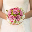 Held close against the intricate lace of a bridal dress, this bouquet appears like a soft cloud of colour and texture, perfectly suited to an elegant ceremony near Golders Green station. At the centre, lush roses in muted lavender-pink tones open fully, their petals thick and cushiony, inviting the fingertips to trace their gentle curves. Tucked between them are bright lime-green button chrysanthemums, compact and spherical, providing a crisp, contemporary contrast that keeps the design feeling fresh and joyful rather than overly traditional. Along the outer edge, creamy white blooms and slim ivory buds circle the arrangement, creating a subtle, ruffled frame that echoes the scalloped hem of a veil. The flowers are bound together in a round, hand-tied style that nestles comfortably in the bride's hands, the stems trimmed evenly and wrapped, likely in a soft ribbon that rests smoothly against her palms. Soft natural light, like that filtering through trees in nearby Golders Hill Park, highlights the interplay between the mauve roses, vivid green pompons, and the white border, giving each element its own quiet moment. The bouquet's imagined fragrance is delicate but present: the comforting sweetness of fresh roses mingling with a hint of green, freshly cut stems. As a composition, it speaks of calm anticipation and heartfelt celebration, the kind of bridal bouquet a local Golders Green florist would craft to look flawless through hours of photos, vows, and evening laughter, holding its shape and colour as steadily as the promises being exchanged.