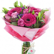 This bouquet radiates an immediate sense of joy and renewal: a hand-tied assembly of vivid fuchsia gerbera daisies with near-black centres, surrounded by softly blushing pink roses at different stages of opening, from tight, elegant buds to full, layered blooms. Small clusters of deep purple-blue statice or similar filler flowers thread through the composition, their papery texture and concentrated colour providing a delicate contrast to the plush petals. Lush green foliage - broad glossy leaves and slender stems - frames each flower, giving the arrangement depth and a fresh, grounded silhouette.
The flowers sit within a clear cylindrical glass vase so the bundled stems are visible below; a length of rustic twine is knotted around the vase neck and a small white rectangular tag hangs from it, printed with a stylised pink-and-green rose logo that identifies the maker. The bouquet is wrapped in layers of sheer, crinkled pink cellophane or tissue that graduates from an almost translucent pastel halo around the blossoms down to a brighter, more saturated pink at the base, catching and scattering soft daylight. The overall scent is gentle and sweet, like an early spring morning in Golders Green when market stalls begin to open - floral, green, and quietly uplifting. As a florist I'd imagine this arranged for birthdays, a thoughtful congratulations, or to lift someone's spirits; its balance of bold gerberas and tender roses makes it equally suitable for lively celebrations and heartfelt, personal gestures across NW11 and nearby Finchley Road.