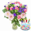 A bright, celebratory bouquet sits in a clear glass vase, photographed against a crisp white background and accompanied by a cheerful cake-shaped Mylar balloon that reads Happy Birthday. The floral composition is lush and full: soft pink roses take centre stage, their velvety petals unfurling from pale blush to a deeper, warm pink, conveying a gentle, romantic feeling. Interspersed among the roses are large pale-pink Gerbera daisies with layered, ray-like petals and buttery yellow-green centres that open the arrangement with a playful, smiling presence. Sprigs of vivid purple filler-likely statice or limonium-are woven generously through the bouquet, their papery clusters adding striking contrast and a textured, almost confetti-like depth against the smooth rose petals. Deep glossy leaves cradle the blooms, offering a rich green backdrop that makes the colours sing and suggests freshness from careful conditioning. The clear vase reveals green stems and water, hinting at recent hand-trimming and attentive care one expects from a neighbourhood florist. To the lower right, the metallic cake balloon is decorated with white 'frosting,' blue piped icing, colorful sprinkles, and four striped candles topped with tiny yellow flames; the multi-coloured Happy Birthday lettering stands out in playful reds, blues, greens and purples, and a small KALEIDOSCOPE mark appears at the very bottom. The overall mood is jubilant and tender, ideal for marking a birthday with both floral beauty and whimsical festivity. A subtle floral scent is suggested-soft, sweet and garden-fresh-making this arrangement feel like a thoughtful, contemporary gift prepared for local celebration in Golders Green and nearby streets.