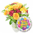 A bright, comforting get-well gift arranged against a clean white backdrop: on the left a full, lush bouquet sits in a clear glass vase, the stems and water visible and lending a simple honesty to the design. Large, softly unfurling roses in warm orange and deep yellow form the heart of the spray, their velvety petals catching a pale daylight that makes the colours glow like a sunny morning in Golders Green. Sunlit yellow gerbera daisies, with their open, golden faces, radiate cheerfulness around the roses while rich burgundy-to-maroon carnations add depth and ruffled texture, a pleasing contrast to the smoother rose petals. Sprays of delicate white alstroemeria peek through the massed blooms, lifting the composition with pale points of light, and abundant glossy green foliage - varied leaves and soft fronds - cradles every stem and gives the bouquet a freshly gathered feel. Slightly overlapping the lower right of the vase is a round, silvery Mylar balloon whose mirror-like surface reflects a hint of the studio light; in its centre a playful cluster of cartoon-style balloons in sky blue, purple, lime green, pink and yellow sport stripes, polka dots and checkerboard patterns. Bold yellow-and-blue letters with a faint shadow spell out Get Well Soon across the balloon, surrounded by tiny starburst motifs along the white rim; a small KALEIDOSCOPE logo sits at the very bottom edge. The overall effect is tender and uplifting - a composition that suggests warmth, scent, and optimism, the kind of thoughtful bouquet one might send from a local Golders Green florist to brighten a recovery at home or in hospital.
