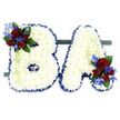 A visually striking and deeply tender floral tribute spells out the capital letters B and A, arranged side by side on a low dark green stand against a clean white backdrop. Each letter is built from a dense, pillowy surface of small pristine white chrysanthemums, their layered petals creating a soft, cloud-like texture that reads as quiet purity and reverence. The contours of both letters are crisply defined by a ruffled royal blue satin ribbon, the rich blue edge giving structure and a solemn, elegant frame to the whiteness. At the upper left curve of the B and the lower right leg of the A delicate accent clusters provide focal points: velvety deep red roses in varied stages from tight bud to gently opening bloom, paired with electric blue delphinium-like spikes that add vertical contrast and a cool, poignant note. Dark green foliage and light green trailing tendrils weave through these accents, softening lines and adding a natural, living depth. Under gentle studio light the white chrysanthemums glow with a diffused, peaceful sheen while the blue ribbon and blooms read as composed and dignified. This composition feels handcrafted by a seasoned florist, balanced for a funeral or memorial where a loved one's initials are celebrated with quiet grace. The scale and stable base suggest a freestanding memorial piece suitable for chapel display or placement beside a casket, conveying respect and personalized remembrance. As a local reflection, this is the kind of bespoke tribute often commissioned by families in Golders Green and delivered near Golders Hill Park or Golders Green station, chosen for its clarity of line, comforting texture, and the clear emotional language of white, red and blue blooms.