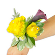 A vibrant, wearable corsage rests diagonally across a light-to-medium forearm against a pristine white background, the composition angled from the lower-left to the middle-right of the image so the arrangement reads clearly as an accessory. Two buttery yellow roses form the heart of the design: one slightly larger and fully open on the left, the other a closer, cupped bud to its right. Each rose carries a small, clear rhinestone nestled near its center, catching the soft studio light and lending a discreet, celebratory sparkle. Slightly behind and above the roses a single calla lily unfurls in a deep plum to near-black cherry tone, its smooth trumpet shape offering a dramatic, sculptural counterpoint to the rounded rose heads. Around these focal blooms a mix of foliage builds texture and depth - feathery asparagus fern provides airy green fronds that halo the flowers, while broader variegated leaves with pale margins (reminiscent of pittosporum) form a stable base beneath the bouquet. Tiny white filler flowers, like miniature spray mums or baby's breath, tuck into crevices to soften edges and enhance fullness. The floral elements are secured to the wrist with a thin band that is mostly concealed, so the flowers appear to sit naturally on the skin; you can almost imagine the faint scent of fresh roses and the cool, waxy feel of the calla's spathe. As a local Golders Green florist's piece, the corsage suggests celebratory uses-prom, a bridal party, or a reception guest-offering a joyful yellow-and-plum palette that reads equally modern and timeless in close-up photography.