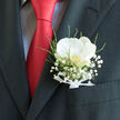 A refined orchid pocket boutonnière sits elegantly in the breast pocket of a dark, finely cut suit, the scene composed as though ready for a sophisticated Golders Green wedding. The focal point is a single white orchid, its petals smooth, velvety, and gently curved, glowing a warm cream under soft, natural light. At the centre, a delicate flush of yellow-gold and pale pink offers subtle depth, like a quiet spark of joy hidden within the flower. Surrounding the orchid, airy clusters of baby's breath create a cloud of dainty white blossoms, adding a dreamy softness and a sense of lightness. Fresh green foliage slips between the blooms, crisp and vibrant, evoking the leafy calm of nearby Hampstead Garden Suburb and the lawns of Golders Hill Park. At the base of the design, a neat white ribbon is tied with disciplined care, the satin edges folded precisely, revealing the practiced hand of a professional florist. The boutonnière rests against a charcoal jacket whose fabric appears slightly textured, providing a strong, structured backdrop that amplifies the flowers' delicacy. A bold red tie cuts through the composition, its rich colour echoing celebration and music-filled evenings in local venues around Golders Green Road. The contrast between the vivid tie, the dark suit, and the pristine bloom creates a sense of poised drama without losing its gentle romance. This small floral piece feels like a quiet declaration of love and commitment, especially fitting for a groom, best man, or proud father on a day when every detail, from the fragrance of the flowers to the shimmer of the ribbon, is chosen with intention and heartfelt sentiment.