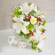 Set against a mellow beige fabric backdrop, this refined table arrangement in a white wicker basket feels like it was designed for a chic Golders Green wedding or intimate family gathering. The basket sits on a spotless white surface, its woven texture providing a subtle, rustic contrast to the elegance of the flowers. Dominating the composition are several large white lilies, their trumpet-shaped blooms gloriously open and lightly speckled, with golden-tipped stamens stretching forward as though offering their fragrance. Just above them, a single cream-to-pale-yellow rose stands as the arrangement's soft heart, its petals wrapped tightly in a classic spiral that hints at romance and new beginnings. Flowing from the right side, white phalaenopsis orchids cascade downward in a gentle waterfall, their rounded petals smooth and luminous, like porcelain butterflies captured mid-flight. Around these central blooms, a lively mix of greenery creates depth and rhythm: feathery grasses, broader green leaves, and bright mossy-green dianthus balls that resemble tiny pom-poms. Threaded throughout are delicate clouds of white baby's breath and clusters of deep red berries, whose glossy surfaces catch the light and add vivid, jewel-like accents. The overall palette of whites, greens, creams, and crimson touches feels both fresh and timeless, ideal for a top table near Hampstead Garden Suburb or a stylish dinner party just off Golders Green Road. To the left of the main basket, two white decorative balls crafted from interlaced strips mirror the wicker's texture and balance the composition, giving the impression of a thoughtfully curated, contemporary centerpiece created with a florist's practiced eye and an appreciation for local Golders Green style.