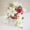 A sophisticated white and red floral display is styled as a luxurious wedding or event table arrangement, captured against a softly textured beige backdrop that recalls the calm interiors of North West London homes. The flowers sit in a round, open-weave white basket that feels both contemporary and artisanal, its delicate latticework allowing glimpses of stems and greenery inside. Rising proudly from this base are large oriental lilies, their pristine white petals open wide and slightly reflexed, with long stamens that cast tiny shadows across their surfaces. Deep red roses, velvety and rich, are tucked among the lilies, their compact blooms adding a passionate, romantic depth that would suit an intimate Golders Green reception or anniversary dinner. Lace-fine sprays of white baby's breath weave through the composition, introducing a dreamy, cloud-like layer that lightens the bold colour contrast. Bright red hypericum berries punctuate the arrangement, nestled among sprigs of light, feathery greenery that gives the whole design a sense of movement and freshness, much like a breeze drifting through nearby Hampstead Garden Suburb. To one side, a graceful arc of white phalaenopsis orchids tumbles gently downward, each blossom delicately formed with hints of creamy yellow and soft pink at its centre, while small buds promise blooms still to come. On the pristine surface beside the main display, a small grouping of white woven spheres echoes the basket's texture, reinforcing the theme of handcrafted elegance. The overall mood is one of timeless celebration and refined style, ideal for wedding tables, engagement parties, or sophisticated festive gatherings across Golders Green and the surrounding area.