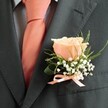 With understated charm, this close-up from Golders Green showcases the art of boutonniere design, celebrating milestone days like weddings and proms in the local community. The focus is on a distinguished suit jacket in slate grey-its tactile luxury brought to life by soft daylight. Beneath, a pale grey-blue shirt quietly supports a satiny peach necktie, its glow reminiscent of warmth on a summer evening. The visual centrepiece is a single peach rose, its form gentle and open, nestling among clusters of airy white gypsophila. Wispy green accents and a broader, richly veined leaf add vitality and depth, framing the flower with natural grace. All is neatly tied with a matching satin ribbon, its peach hue uniting the boutonniere and tie in an effortless harmony. There's an ambience of anticipation-a breath of fresh blooms, a hint of delicate fragrance, and the subtle thrill of a special event unfolding on the avenue. Crafted with sensitivity to both tradition and the local spirit, this boutonniere mirrors the love, care, and unity that hallmarks celebrations in Golders Green.