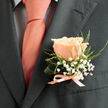 Captured in gentle, directional light, this intimate close-up shows a single peachy-pink rose boutonnière resting against a deep charcoal suit lapel, a study in restrained elegance. The central rose is just opening, its petals layered with subtle gradations from blush to warm peach, each edge catching light and revealing velvety texture. Surrounding the flower are delicate sprays of white gypsophila whose minute blooms create a soft, cloud-like halo, while fine green sprigs add a whisper of organic shape and a cool contrast to the suit's structured lines. A pale pink satin ribbon, narrow and lustrous, is tied at the base of the stems, its sheen harmonising with the warmth of a matching tie and the faint, dark-grid pattern of a light-coloured shirt beneath the jacket.

The composition balances softness with sartorial formality: the rose's tactile warmth and the gypsophila's airy lightness temper the sharpness of tailored wool, making the accessory feel personal rather than ornamental. One can almost sense the bloom's delicate scent and the quiet ceremony of fastening a boutonnière before leaving home - perhaps for a wedding near Golders Hill Park or a celebration after a service at a local chapel. This handcrafted piece reflects the practiced touch of a neighbourhood florist who understands how small botanical choices - colour tone, scale, texture - translate into a meaningful accent on an important day in Golders Green and the surrounding streets.