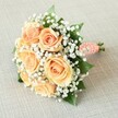 Vividly capturing the celebratory atmosphere of a Golders Green event, this elegant bouquet lies diagonally across a pale, lightly textured linen surface, its rounded shape drawing the eye. Dominated by seven to eight full, peach-hued roses, each flower radiates warmth akin to a soft summer evening in Hampstead Garden Suburb. The roses' petals delicately spiral outward, creating a dynamic interplay of apricot and gentle orange tones. Scattered among the roses, tiny starbursts of baby's breath form airy clusters reminiscent of morning dew over Golders Green Crematorium gardens, their crisp whiteness contrasting beautifully against the roses. Slim, lustrous green leaves peek out, anchoring the bouquet and echoing the verdant scenery of local parks. The stems are expertly gathered and secured with a peachy satin ribbon, accented with a graceful twist of pearly beads-alluding to the understated elegance found in local Golders Green ceremonies. A touch of the freshly cut stems remains visible at the base, reinforcing the bouquet's natural beauty. Soft, diffused lighting skims across the petals, bringing out their gentle textures and subtle sheen. This bouquet, designed with care by skilled Golders Green florists, conveys romance, freshness, and an unwavering sense of joy, making it a heartfelt addition to weddings and special occasions throughout the neighbourhood.