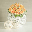 This composition reads like a lesson in understated elegance: a tightly domed cluster of peach and apricot roses, lavish and full, rests atop a sculptural white woven orb whose open lattice gives the arrangement an airy, contemporary lift. Clouds of white gypsophila weave between the roses, creating a frothy veil that brightens the peach tones and scatters soft highlights across the petals. Lush foliage frames the base, adding cool, verdant texture and a subtle contrast to the warm blooms. The light in the image feels like late-morning glow from Golders Green Road, falling gently across each curl of petal and the wicker strands, so that the whole piece seems to breathe with a soft, luminous calm. You can almost sense the faint, sweet perfume of the roses mingling with the green, an inviting, powdery aroma that suits weddings, milestone anniversaries, or thoughtful gifts for someone moving into a new flat near the Tube. As a local florist with roots in the neighbourhood, I notice the composition's practical beauty-the stable spherical vase for transport, the airy structure that reads well on a mantel or as a centrepiece-and the care shown in the placement of every bloom. Flowers Golders Green prepares pieces like this for customers across NW11, often chosen for their quiet sophistication and ability to suit both contemporary interiors and traditional celebrations. The bouquet's tactile contrasts-the soft velvet of petals, the whisper of baby's breath, the crisp weave of rattan-make it not just seen but felt, a sensory expression of thoughtful craftsmanship in the heart of Golders Green.