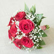A lush cluster of deep crimson roses and airy white baby's breath lies carefully arranged on a light, textured fabric, like a romantic still life captured somewhere just off Finchley Road in Golders Green. The bouquet is tightly composed, with around six or seven large red roses at its core, each bloom fully open yet still structured, showing layers of velvety petals that curl and fold like soft satin. Nestled between these ruby centres, intricate stems of gypsophila form frothy clouds of tiny white blossoms, their fine stems and starry flowers filling every gap so there is no harsh line, only a gentle transition from bold colour to soft lightness. Encircling the flowers, a fan of broad, dark green foliage provides contrast and volume, the leaves' smooth, slightly glossy surfaces catching the light and grounding the arrangement in natural calm, reminiscent of greenery from nearby Hampstead Heath. The stems are bundled together in a compact, hand-held style and wrapped with a crimson ribbon that matches the roses, the fabric wound neatly around the handle and decorated with a subtle spiral of white bead-like dots. This small detail adds a touch of bridal refinement, as though the bouquet is waiting to be carried down an aisle at a local Golders Green ceremony or presented as a heartfelt romantic gesture on a special evening. The overall impression is one of careful craftsmanship, balance, and deep affection, making it ideal for weddings, anniversaries, or any moment when love needs a classic, elegant voice.