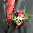 This image captures a carefully crafted boutonnière, the sort of intimate floral detail a local Golders Green groom might choose for a ceremony in a nearby synagogue or a chic registry office celebration. The design centres on a beautifully formed hot pink rose, its petals unfurling in smooth, velvety layers that curl protectively around the core, as though sheltering a treasured secret. Surrounding the rose is a delicate collar of white baby's breath, each miniature flower forming a cloud-like ring that softens the strong pink and suggests purity, tenderness, and enduring affection. Fine, feathery strands of decorative grass arc around the arrangement, adding a whisper of movement and a hint of wildness, reminiscent of the gentle breeze that passes through the open spaces near Hampstead Garden Suburb. A single, deep green leaf, slightly waxy and reflective, supports the whole piece from behind, providing depth and contrast so the rose can truly stand out. At the base, a small red ribbon is tied into a tidy bow, its satin finish catching the light and echoing the romantic theme without overpowering the flowers. The boutonnière rests against a dark, fitted suit, the rich neutral cloth working like a stage curtain that lets the colours shine. As a whole, the piece feels fresh, intimate, and thoughtfully composed, ideal for a wedding day, engagement party, or milestone celebration where every detail matters and where flowers, like those prepared by Flowers Golders Green, help tell the story of love and commitment.