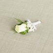 A classic white rose boutonniere lies diagonally across a softly textured, light beige fabric that resembles natural linen, the gentle crosshatch weave echoing the understated elegance of a North West London wedding morning. At the heart of the design is a single creamy white rosebud, its velvety petals still tightly furled yet full and promising, with a faint hint of green at the base where the sepals cradle the bloom. Just behind and to the side of the rose, tiny white clusters of baby's breath float like miniature clouds, their airy flowers softening the outline and giving the piece a romantic, almost ethereal lightness. Slender, deep green leaves and a few feathery sprigs of foliage fan out discreetly, adding depth, contrast, and a fresh, garden-picked feel, as if gathered from a quiet corner near Golders Hill Park. The stems are neatly wrapped in a pristine white satin ribbon, smooth and slightly lustrous, meticulously coiled around the base and finished with a delicate, proportionate bow that sits just below the flower head, ready to be pinned to a lapel. Soft, diffused lighting washes over the boutonniere, catching the gentle curves of each petal, the tiny buds of gypsophila, and the sheen of the ribbon without casting harsh shadows. The overall impression is one of purity, tradition, and quiet sophistication, a timeless accessory suited to a groom or groomsman arriving at a ceremony in Golders Green, Hampstead Garden Suburb, or along Finchley Road, capturing the emotions of new beginnings, commitment, and discreet, refined style.
