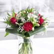 A richly detailed red and white bouquet stands proudly in a clear cylindrical glass vase, its freshly cut green stems visible beneath the waterline like a tidy forest of colour and life. At the heart of the arrangement are velvety deep red roses, their petals just beginning to unfurl in spirals that suggest timeless romance and quiet devotion. Nestled beside them, a large ruffled crimson carnation adds intricate texture, its many layers catching the soft daylight that seems to stream in as if from a Golders Green townhouse window. Threaded through this passion-red centre are pure white and creamy blooms: star-shaped white alstroemeria with fine speckles at their throats, plump white chrysanthemum heads, and several closed white lily buds poised to open over the coming days. Tiny clusters of baby's breath float like pale mist between the larger flowers, softening the contrasts and giving the bouquet a gentle, cloud-like fullness. Around the base, glossy dark green tropical leaves are carefully curled and wrapped, forming an elegant collar that mirrors the tidy pavements near Golders Hill Park. Eucalyptus-style sprigs and pointed green foliage stretch upwards, adding layers of green in cool and warm tones, from silvery sage to deep forest. The bouquet rests on a clean white surface against a softly blurred, bright background, suggesting a calm interior space in Golders Green ready to welcome guests. Crafted with care, this floral display feels ideal for anniversaries, milestone birthdays, Mother's Day, or to bring a touch of polished celebration to a local office or home, its colours speaking of love, gratitude, and heartfelt warmth.