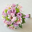 Placed thoughtfully on a softly textured neutral surface, this radiant hand-tied bouquet glows with a harmonious medley of pink, lavender, lime green, and white-an uplifting expression of joy from the heart of Golders Green. The bouquet's core features full-bloomed lavender roses whose tender spiraled petals evoke quiet elegance and romance, reminiscent of Golders Green's welcoming community spirit. Interlaced throughout are alstroemeria lilies in pale pink, each blossom brushed with yellow and dark pink, lending both subtle warmth and artistic flair. Clustering between the pastels, bright lime green button chrysanthemums provide lively contrast and a modern edge, their beadlike shapes catching the light hinting at contemporary gatherings around Finchley Road. Wisps of white baby's breath create a gossamer softness, filling the spaces with ethereal delicacy, while glossy green leaves peek between the flowers, grounding the design in freshness and vitality. The composition is brought together by a fuchsia satin ribbon, tastefully wound around the neatly trimmed stems and embellished with iridescent pearl beads-a romantic detail that nods to wedding celebrations and anniversaries across NW11. This bouquet is an inviting centerpiece suited for joyful milestones, reflecting artistry and freshness that only a knowledgeable Golders Green florist can provide.