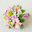 A romantic bouquet lies diagonally across a gentle, oatmeal-toned fabric background, the kind of natural, woven texture that lets the flowers take centre stage. From this slightly overhead view, layers of soft pink roses unfurl in perfect spirals, their petals overlapping like silk, creating a sense of calm assurance and timeless love that would suit an intimate ceremony near Golders Green station or a quiet celebration at home. Between them, bright lime-green button chrysanthemums form neat, rounded domes, each tiny petal catching the light and bringing a fresh, modern pop of colour that keeps the design lively rather than overly traditional. Pink alstroemeria, with their slender, arching petals, introduce a note of subtle drama: blush and rose tones are brushed with deeper pink streaks and flecks of warm yellow, giving the impression of fine watercolour work viewed close-up on a florist's bench near Temple Fortune. Wisps of white baby's breath drift between the larger blooms, adding a soft, misty layer that lightens the overall look and lends a fairy-tale quality, like a veil of tiny stars. Around the edges, cool green foliage fans out, adding structure and a garden-fresh character, as though the stems were gathered from a hidden corner of Hampstead Heath. The stems themselves are gathered tightly and bound in a satin magenta ribbon, the sheen of the fabric catching the light and reinforcing the bouquet's celebratory purpose. Everything is lit with gentle, even brightness, revealing texture without harsh shadows, making this arrangement feel like a promise of joy ready for wedding delivery across Golders Green and the surrounding North West London neighbourhoods.
