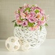 On a pristine white tabletop, a generous floral centerpiece overflows from a sculptural white woven vase, its open latticework catching the soft studio light like delicate basketry. The rounded container, crafted from slim white strips that criss-cross and twist around each other, feels both contemporary and a little rustic, the kind of piece you might spot in a stylish Golders Green living room overlooking Hampstead Garden Suburb. Rising from this textured base is a lush, dome-shaped bouquet of fresh flowers, dominated by fully opened roses in pastel pink and gentle lavender, their velvety petals layered tightly in spirals that suggest romance and quiet celebration. Threaded among the roses are pink alstroemeria, the Peruvian lilies marked with fine stripes and freckles that draw the eye into their trumpet-shaped blooms, some showing hints of soft yellow or deeper rose tones at the throat. Bright lime green button chrysanthemums nestle between the petals like tiny cushions, their compact, pom-pom heads adding playful contrast and modern flair. Sprigs of airy white baby's breath scatter light, cloud-like clusters throughout the bouquet, softening the colour transitions and giving the arrangement a dreamy, almost floating quality. Glossy dark green leaves peek out here and there, grounding the pastel palette with a touch of natural depth. To the left of the vase, two decorative woven spheres in pale, natural tones echo the vase's texture, one larger and one smaller, arranged slightly off-centre for visual balance. Behind everything, a textured light beige wall recedes quietly, allowing the pinks, lavenders, and limes to glow, like a thoughtfully styled centerpiece ready for same-day delivery to a Golders Green dinner table or intimate birthday gathering.
