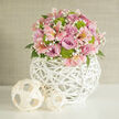 This image shows a beautifully composed, rounded floral arrangement that seems to glow with quiet joy, much like a calm afternoon wandering through the leafy streets around Golders Green station. Soft lavender and rosy pink roses form the main body of the bouquet, each bloom carefully opened to reveal velvety petals arranged in concentric curls. Nestled among them are pale pink alstroemeria, sometimes called Peruvian lilies, their long petals brushed with subtle freckles and stripes that add an element of playfulness and movement. Clustered tufts of vivid lime green button chrysanthemums pop out from the softer tones, giving the design a crisp, contemporary edge and adding texture you can almost feel with your fingertips. Throughout the arrangement, airy sprigs of tiny white baby's breath weave in and out, creating a light veil over the larger blossoms. Dark green leaves tuck underneath and occasionally peek through, framing the flowers with natural depth. The entire bouquet is nestled in a round, white woven vase made from thin wicker or rattan strips, arranged in an open, basket-like lattice that looks modern yet organic. This sculptural globe rests on a pristine white surface, which subtly reflects the light and echoes the brightness of the vase. To one side, two smaller decorative balls made of pale woven material sit casually, tying together the natural textures. Behind, a softly textured beige backdrop keeps the focus entirely on the flowers. The overall feeling is calm, fresh, and quietly celebratory, perfect for a wedding collection display or as a romantic centrepiece in a Golders Green home.
