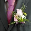 A close-up photograph focuses on the upper torso of a man dressed for a special occasion in Golders Green, showing only his charcoal grey suit, pale patterned shirt, and a vivid magenta-pink silk tie neatly knotted at his throat. Pinned to the left lapel, just above the fold of the crisp jacket, is an exquisitely crafted boutonniere that immediately draws the eye. At its heart sits a delicate light pink rose, its petals softly unfurling in layered spirals that look velvety to the touch, the sort of bloom you might see freshly chosen from a florist near Golders Green Road on a wedding morning. Nestled beside the rose is a smaller pink flower, reminiscent of an alstroemeria, its inner petals streaked with deeper purple-pink markings that add a painterly detail. Small clouds of white gypsophila cluster around the main flowers, their tiny blossoms creating a halo of softness and innocence, like a fine mist. A compact green button chrysanthemum adds a fresh pop of lime colour, giving the design a modern, almost playful contrast against the pastel tones. Feather-light fern fronds and slender blades of greenery arc around the blooms, providing movement, depth, and a slightly woodland feel, as if the flowers were gathered from nearby Hampstead Heath. The stems are carefully bound with a narrow lavender satin ribbon, tied into a neat bow that shimmers subtly against the suit fabric and gently echoes the pink of the tie. The overall impression is one of elegant celebration, capturing the quiet excitement of a wedding or prom evening somewhere in north-west London, ready to accompany the wearer through moments of vows, toasts, and photographs with family and friends.