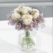 Close-up image of an elegant white and lavender bouquet arranged in a textured glass vase, styled as if it has just been set down on a light, modern table in a calm Golders Green home. At the heart of the design, several creamy white roses open in full, their velvety petals gently spiralling out from centres touched with the faintest hint of buttery yellow, giving a feeling of freshness and quiet luxury. Around these classic blooms, clouds of tiny lavender-purple flowers, likely statice or limonium, create a delicate halo, their papery, clustered petals forming a soft haze that contrasts beautifully with the smooth, substantial rose heads. The stems, a clear healthy green, are visible through the water in the vase, grounding the arrangement and emphasising its just-cut vitality, as though gathered that morning from a nearby market stall off Golders Green Road. The vase itself has a distinctive crackle, almost ice-like texture that catches the natural light and scatters it into gentle highlights, adding a subtle sparkle without distracting from the flowers. The bouquet rests on a bright white surface that reflects the light, casting faint, feathery shadows beneath the vase and contributing to a serene, uncluttered atmosphere. Behind, the background melts into a soft, pale grey blur, keeping every bit of attention on the play of colour and form in the flowers. The overall mood is tranquil and comforting, the kind of arrangement that might sit on a bedside table in a quiet flat near Hampstead Garden Suburb, or on a reception desk, offering understated sympathy or a thoughtful get-well gesture with timeless, soothing grace.