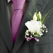 Pinned carefully to the lapel of a crisp charcoal jacket, this wedding boutonnière feels like a quiet love letter carried close to the heart, the sort of refined detail you might see as couples stroll from Golders Green station towards a synagogue or reception nearby. The focal point is a fully opened white lisianthus, its petals softly frilled and delicately layered, almost translucent at the edges where the light grazes them. Just behind, two slender buds stand upright, their green tips still folded, symbolising new beginnings and the tender anticipation of married life. Wisps of baby's breath surround the lisianthus, forming a light halo of tiny, star-like blossoms that soften the contrast against the dark suit fabric. Slender strands of green foliage thread through the design, bringing a sense of movement and natural spontaneity, while a single rich green leaf is curled into a partial loop beneath, offering structure and depth. A narrow mauve ribbon, tied into a gentle bow at the stem, introduces a hint of romantic colour that quietly harmonises with the groom's silk tie, catching a muted sheen when the light hits. The arrangement looks freshly crafted by a local Golders Green florist, with the blooms holding a faint, clean fragrance that seems to follow the groom as he greets guests and shares smiles. The overall impression is one of understated sophistication and heartfelt celebration, a small but meaningful floral accent marking a day filled with vows, family, and enduring connection.