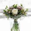 An elegant white and green bouquet is captured in soft daylight, standing in a simple clear glass vase against a luminous, blurred white background that feels like a bright Golders Green window on a quiet morning. At the heart of the arrangement, three creamy white roses are fully open, their velvety petals gently spiralling around subtle ivory centres that suggest absolute freshness and purity. Around them, a collar of lush green foliage cradles the blooms, with broad, slightly folded leaves in deep emerald tones giving the bouquet structure and a polished, professional finish. Threaded among the roses are delicate sprays of white gypsophila, or baby's breath, their tiny blossoms floating like a fine mist and adding a light, cloud-like texture that softens the whole design. Slender stems of alstroemeria, some still in bud and others just beginning to unfurl, introduce gentle hints of muted purple and pale blush-pink, their streaked petals promising more colour as the days pass. The flowers are arranged in a classic, rounded dome, neat and compact without feeling crowded, the stems clearly visible through the cylindrical glass vase, gathered in a tidy bundle and secured just above the waterline with a discreet ribbon. The clear water glints in the natural light, reinforcing the sense of just-cut freshness you might expect from a trusted local florist. The overall mood is calm and refined, ideal for a thoughtful gesture of comfort, a quiet celebration, or a serene wedding bouquet delivered to a home near Golders Hill Park or a peaceful corner off Finchley Road, bringing a sense of gentle, timeless beauty into the room.