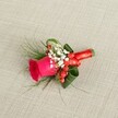 A richly detailed buttonhole arrangement lies against a softly textured, light beige linen-style fabric, the neutral background allowing every colour and curve to stand out clearly. At the heart of the design sits a deep fuchsia pink rose, its velvety petals tightly layered in the centre and gently unfurling towards the outer edges, giving a sense of freshness and quiet luxury. Clusters of tiny white baby's breath blossoms nestle at the base of the rose, their cloud-like texture adding a delicate, romantic haze that softens the boldness of the central bloom. Threaded between these white flowers are small, glossy red hypericum berries, their rounded shapes catching the light and adding a lively, festive accent that suggests celebration and joy. Surrounding the rose is a collar of rich greenery: some leaves are broad and a darker, polished green, carefully curved and folded to cradle the flowers, while finer, feathery sprigs of fern-like foliage extend outward with a light, airy movement. This gentle mix of textures feels like something you might see carried down a quiet Golders Green side street on the way to a ceremony near Hampstead Garden Suburb. The stems are bound tightly and neatly with a vivid red satin ribbon, wrapped with a practised florist's touch and finished in a small, elegant knot or bow that reinforces the romantic, formal mood. The soft, even lighting reveals every detail without harsh shadows, making this boutonniere ideal for a groom, best man, or prom guest wanting a refined yet vibrant floral accent for a special day in and around Golders Green.