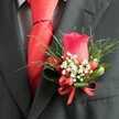 A close-up, high-angle photograph reveals a meticulously crafted buttonhole pinned to the left lapel of a deep charcoal suit, the kind you might see on a groom stepping out of a car on Finchley Road in Golders Green. At the heart of the arrangement stands a single, velvety rosebud in a rich, deep red, its petals tightly coiled yet full of promise, catching the soft indoor light. Clusters of tiny white flowers, reminiscent of baby's breath, nestle at the base of the rose, their airy, cloud-like texture softening the strong lines of the suit fabric. Glossy red berries, oval and polished, peek through the white blooms, echoing the colour of the rose and adding a subtle sense of movement and depth. Surrounding everything is a frame of lush greenery: slender, grass-like fronds fan out delicately, while broader, darker green leaves are wrapped securely around the base, giving structure and balance. A neat red satin ribbon is tied into a small bow around the stems, its sheen mirroring the smooth, bright red tie visible beneath the lapel, perfectly knotted against a finely patterned light grey or white shirt. The overall effect is one of refined elegance and careful coordination, an intimate portrait of formal menswear accented by thoughtful floristry. You can almost imagine this boutonnière being adjusted just before a wedding at a local synagogue or a celebration near Golders Hill Park, the fresh scent of greenery and the quiet confidence of the wearer combining to mark a significant, joy-filled moment.