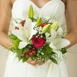 Lush with intention and grace, this bridal bouquet evokes the inviting gardens of Golders Green: radiant white lilies unfurl broadly, their glossy petals a testament to purity and joyous beginnings. Close by, deep crimson roses glow with velvety richness, each tight spiral reflecting the enduring flame of romance. The bride's white, subtly patterned gown frames the composition, her hands protectively intertwined around the bouquet-a gesture full of serenity and hope. Tiny clouds of baby's breath drift throughout the bouquet, their delicacy providing a soft, romantic haze that evokes mornings along Golders Green Road. Pops of ruby red hypericum berries glisten among unique round clusters of mossy green, likely dianthus or related foliage, offering whimsical textural surprises. Broad green leaves and feathery ferns form a lush base, their varied hues mirroring the natural tapestry of Golders Hill Park just nearby. The bouquet, crafted with a meticulous attention to local favourites and seasonal freshness, is imbued with the gentle excitement of a wedding day in NW11. Every detail, from closed lily buds to the soft light brushing the bride's hands, is chosen to celebrate both personal memories and the timeless beauty of Golders Green-a bridal bouquet meant to linger in the heart long after vows are spoken.