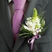 A close-up, high-angle photograph captures the upper torso of a man dressed for a formal occasion in Golders Green, his attire centred around a deep charcoal grey suit jacket. The lapel is sharply tailored, lying flat and smooth, with the stitching and clean shoulder line hinting at careful preparation for a wedding or smart celebration. Beneath the jacket, a pale dress shirt with a faint grid pattern peeks through, its cool tone perfectly offset by a sleek lavender tie that drops in a clean line down the chest. Pinned to the left lapel is a meticulously crafted buttonhole, the clear star of the image. At its highest point rises a narrow spire of pale green buds, likely hyacinth or Muscari, still tightly closed and full of promise. Around this, open-faced white alstroemeria blooms unfurl, their petals brushed with fine maroon and plum streaks that read like hand-painted markings, golden stamens glowing softly at their centres. Tiny white clouds of gypsophila weave between the larger flowers, the minuscule blossoms adding a hazy, romantic softness. Slender grass-like blades and darker, broader green leaves arc and nestle around the florals, adding structure and a fresh, garden-picked feel reminiscent of nearby Golders Hill Park on a spring morning. At the base of the buttonhole, a neat satin ribbon in lavender, perfectly matched to the tie, is tied in a small, precise bow, holding the stems and foliage in a compact cluster. The overall effect is one of quiet sophistication and understated romance, a groom or groomsman in Golders Green ready to step into a day filled with promise and celebration.