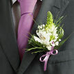 This close-up image captures the refined details of a man's formal attire, perfect for a Golders Green wedding, prom, or black-tie celebration along Finchley Road. The focus is on the left lapel of a deep charcoal grey suit jacket, its smooth weave and careful stitching hinting at quality tailoring, with a sharp lapel edge that naturally draws the eye to the floral accent. Underneath, a pale shirt with a fine dark check or gingham pattern adds understated character, while a sleek lavender necktie, tied in a classic knot, flows down the centre of the frame. The tie's soft lilac sheen is mirrored in the satin ribbon of the boutonniere, binding the whole look together. The boutonniere itself is a delicate composition of white Peruvian lilies, their open petals marked with fine reddish-brown streaks and golden-yellow centres that suggest gentle fragrance and warmth. Rising proudly from the heart of the arrangement is a tall, conical green bud, probably tuberose, its lime-green tone bringing fresh contrast and vertical interest. Around it, tufts of tiny white gypsophila blossoms form a cloud-like halo, giving the piece a light, romantic air. Various stems of greenery, from fine, spiky blades and feathery textures to broader deep green leaves, create structure, depth, and a lush backdrop to the pale blooms. At the base, a lilac satin ribbon is tied into a neat bow, its loops and tails lying smoothly against the lapel as it secures the stems. The overall mood is calm, confident, and hopeful, as if the wearer is standing just outside a synagogue or reception venue in Golders Green, ready to step into a cherished moment.