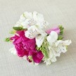 A compact, hand-tied floral arrangement lies horizontally on a softly textured, light beige fabric that looks like natural linen, the neutral background gently cradling the colours. At the heart of the design is a lush fuchsia-magenta bloom, peony-like in its fullness, with layers upon layers of ruffled petals that seem almost velvety to the touch. Around this central flower, several white alstroemeria, or Peruvian lilies, open gracefully, each trumpet-shaped bloom marked with fine maroon and burgundy streaks and freckles that draw the eye inward, as if sketched with a delicate pen. Slim, elongated green buds and spiky florets are woven between the blossoms, adding fresh, spring-green accents and a slightly linear texture that contrasts with the softness of the petals. The stems are neatly gathered and wrapped in a bright pink satin ribbon, gleaming gently and echoing the vivid hue of the central flower, giving the piece a polished, modern finish. The lighting is soft and even, with gentle shadows that suggest calm indoor daylight, highlighting the freshness of the flowers without any harsh glare. The arrangement feels like a small corsage or boutonnière created for a wedding or prom, something you might see being carefully pinned before a celebration at a Golders Green venue or on the way to an event in nearby Hampstead Garden Suburb. It carries a mood of joyful romance and intimate festivity, as if crafted by a local florist who understands how to balance vivid colour with quiet elegance for special occasions across NW11.