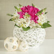 A compact, sculptural arrangement rests on a clean white surface, its design poised between modern minimalism and romantic charm, like something curated for an intimate Golders Green wedding reception. The eye is first drawn to a cluster of rich fuchsia blooms-plush peonies and similarly ruffled flowers gathered closely at the centre-each petal layered and slightly curled, creating a feeling of lush abundance. Encircling this vivid heart, ivory alstroemeria form a soft collar, their slender petals opening gracefully and catching the light with a gentle sheen. Fine speckles and faint markings add delicate detail to the alstroemeria, giving the bouquet a hand-painted quality. Fresh green leaves and slender stems weave through the composition, while tiny unopened buds rise here and there, adding a sense of movement and promise, as though new blossoms could unfurl at any moment. The flowers are nestled firmly into a round, bright white wicker vessel whose intricate latticework feels both contemporary and classic, reminiscent of the artisanal touches found in small shops around Golders Green Road. Nearby, two creamy woven spheres sit quietly, echoing the basket's texture and providing visual balance, like understated décor pieces on a wedding table. Soft, diffused light bathes the scene, enhancing the fuchsia tones, brightening the whites, and deepening the greens, until the whole arrangement seems to glow with understated elegance. It's easy to imagine this piece as a centrepiece for a bridal brunch in a home off Hoop Lane, or as a thoughtful congratulatory gift delivered across NW11. The mood is serene yet joyful-an ode to fresh beginnings, heartfelt gratitude, and the pleasure of beautifully arranged flowers.