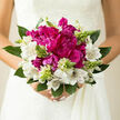 In this photograph, a bridal bouquet is held close, the flowers gathered in a way that feels intimate and composed, as if the bride has just stepped out of a quiet ceremony in Golders Green. Central to the design are sumptuous magenta peonies, each head round and deeply cupped, their ruffled petals layered so generously that they appear almost cloud-like. Around them, white alstroemeria open in star-shaped blooms, their petals slim and slightly curved, each one etched with fine, painterly streaks in soft mauve and plum, bringing a subtle intricacy that invites a lingering gaze. Nestled between these main flowers are upright spikes of pale green buds, some clustered, some more solitary, creating movement that draws the eye gently outward. The foliage is clean and purposeful, with broad, cool-toned leaves forming a natural frame that grounds the arrangement without adding heaviness. The lighting is soft and natural, reminiscent of an overcast yet luminous afternoon above the tree line in nearby Hampstead Garden Suburb; shadows are gentle, and the colours appear true and calm. The bouquet suggests a light, fresh fragrance-green, slightly dewy, with a whisper of floral sweetness-as though the stems have just been conditioned in a local Golders Green studio moments before delivery. The overall mood is one of modern elegance with a classic heart, a design as suited to a chic reception near Golders Green station as it would be to an intimate celebration at home. It feels like a bouquet made for someone who values quiet beauty, careful craftsmanship, and the emotional weight of each flower chosen.