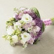 An opulent hand-tied bouquet rests gracefully on a soft beige, subtly textured cloth-its colors and textures a nod to the refined tastes found throughout Golders Green. At the core, multiple white lisianthus blooms open beautifully, showcasing creamy yellow hearts framed by soft, cloud-like petals and accompanied by several delicate, unopened buds. Pastel clusters of light pink and lavender-hued stock flowers weave throughout the bouquet, their ruffled forms offering volume and an invitingly tactile experience. The visual drama intensifies with spherical heads of deep purple Allium 'Star of Persia,' their spiky florets emerging confidently among the lighter florals to provide striking color contrast and dynamic structure. Intermingling tendrils of slender green foliage and bright green lisianthus stems enhance the sense of freshness, reminiscent of the manicured gardens and avenues that shape the Golders Green landscape. All stems are expertly gathered and secured with a vivid fuchsia pink satin ribbon, adorned along its length with an elegant string of white pearls, a finishing flourish synonymous with both romance and sophistication for weddings and special occasions. Bathed in soft, natural light, every hue and detail is lovingly revealed, making this arrangement an irresistible choice for bridal bouquets, elegant event centerpieces, or bringing heartfelt celebration to the heart of Golders Green, where local florists take immense pride in their craft and community.