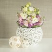 A serene and meticulously crafted floral arrangement sits atop a luminous white tabletop, capturing the natural grace and fresh energy of Golders Green. At its core lies a spherical vase, fashioned from intricately looped strands of white-painted rattan, lending an airy, modern touch that echoes the creative spirit of NW11's artisans. Bursting from the vase are abundant creamy white and buttery yellow eustoma-also known as lisianthus-each bloom soft and ruffled like the early spring blooms one might spot in Golders Hill Park. Wisps of blush pink stock flowers rise among the eustoma, subtly perfumed and adding delicate height. To ground the display, clusters of vivid purple filler flowers-possibly aster or small allium-are set among glossy, deep green foliage, offering rich contrast and texture. Alongside the vase, two decorative open-work spheres made from pale rattan reinforce the airy, handmade theme; their woven patterns play with light and shadow, further enhancing the composition's tactile appeal. The arrangement stands before a softly textured beige wall-warm and inviting, much like Golders Green itself. Together, these elements convey a sense of renewal and quiet celebration, perfect for occasions ranging from wedding breakfast tables to cherished at-home gatherings, promising a lasting impression crafted by local floral experts.