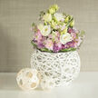 On a smooth white surface, a carefully composed floral arrangement stands in tranquil balance, as if bringing a little bit of Golders Green's soft morning light indoors. The bouquet is rounded and full, centred around ruffled lisianthus blossoms in creamy white and buttery pale yellow, each bloom unfurling in generous, velvety layers. Tucked between them are scented spikes of blush pink stock and clusters of small, spiky lavender allium-style flowers, whose cooler purple notes lend a gentle contrast to the warmer pastels. Slender stems of fresh greenery weave through the design, while a darker green band of leaves encircles the base of the bouquet, neatly gathering the stems together. All of this rises from a distinctive spherical vase made of thin, white branches, crisscrossed and intertwined into a hollow, woven orb. The latticework feels airy and sculptural, allowing just enough space to glimpse the stems inside, much like looking through tree branches in nearby Golders Hill Park. Beside the vase, two natural wicker balls-one slightly larger than the other-rest casually on the tabletop, their pale wooden strips echoing the organic texture and circular shape of the vessel. The background is softly blurred and textured, a warm beige wall that suggests a calm living room or boutique hotel lobby off the high street. Light falls evenly across the scene, highlighting the crisp whites, pastel florals, and tactile materials. The overall impression is of gentle sophistication and quiet reassurance, perfectly suited to a bedside table, a welcome-home gift after a long day commuting through North West London, or a thoughtful floral accent for a soothing, contemporary space.