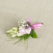 A delicately crafted floral corsage or boutonniere rests on a softly textured, light beige linen background, the woven fabric resembling the understated elegance of a well-laid table in a Golders Green townhouse. At the heart of the piece, a compact cluster of ruffled, soft pink blooms-reminiscent of miniature stocks or dainty garden flowers-unfolds in layers, each petal carrying a gentle gradient from blush at the centre to a slightly paler edge. Threaded between these pastel petals, tiny white baby's breath create a cloud-like halo, their airy heads adding lightness and a sense of movement, as though caught in a quiet breath of air. Here and there, fine sprigs of small lavender or deep purple florets peer out, adding depth and a whisper of evening colour that enriches the pink-and-white palette. To one side, several creamy white to pale green buds, likely freesias or similar slender blooms, stretch gracefully away from the main cluster, still closed and full of promise, suggesting the anticipation of a special day-perhaps a wedding at a nearby Golders Green synagogue or a celebration on Finchley Road. A base of lush, broad emerald leaves grounds the design, their smooth surfaces catching the light, while finer, feathery green foliage extends in wisps from beneath, giving the accessory a slightly wild, garden-fresh character. Around the neatly bound stems, a vivid fuchsia satin ribbon is wrapped with care, tied into an elegant bow whose satin tails lie softly against the linen, adding a celebratory flash of colour. The overall impression is of freshness, tenderness, and thoughtful craftsmanship, a refined floral accent perfectly suited to a lapel, wrist, or clutch bag for any formal gathering in and around NW11.