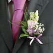 A close-up photograph focuses on the upper body of a man in formal dress, likely a groom standing ready for his ceremony in Golders Green. He wears a dark charcoal suit jacket with a finely textured lapel, layered over a crisp, pale shirt patterned with a subtle grid. At his throat sits a vibrant magenta silk tie, perfectly knotted and catching the soft, natural light with a gentle sheen. Pinned neatly to his left lapel is a meticulously crafted buttonhole: slender ivory buds, reminiscent of snapdragons or lisianthus, rise gracefully at the top, while below them cluster soft pink and lavender blooms that echo the romantic colours often chosen for North London weddings. Tiny clouds of white gypsophila tuck in between the petals, their airy sprays adding a delicate, almost mist-like softness, while a few deeper purple florets bring depth and contrast. Broad, glossy dark green leaves form a supportive base, their smooth surfaces catching hints of light, and feathery, brighter green foliage fans out lightly, giving the arrangement movement and a garden-fresh feel, as if just gathered from nearby Golders Hill Park. At the base of the stems, a slim satin ribbon in lavender is tied into a neat bow, its gentle shine complementing the tie and linking the colours together with quiet elegance. The overall effect is one of poised sophistication and joyful anticipation, the kind of thoughtfully coordinated buttonhole a local florist near Golders Green station might prepare for a spring or summer wedding, marking the wearer as someone cherished on a day of celebration.