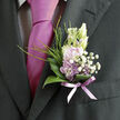 This close-up of a boutonnière resting against a dark, tailored suit jacket captures a moment of gentle elegance, the kind often seen at intimate weddings around Golders Green and nearby Hampstead Garden Suburb. At the centre sits a cluster of lilac and pale lavender blossoms, each petal soft and slightly curved, like a whisper of colour against the charcoal fabric. Surrounding them, tiny white baby's breath forms a halo of airy texture, its miniature blooms creating a sense of lightness and romance, as if a spring breeze had been gathered and pinned in place. Smooth, glossy green leaves frame the arrangement, arching protectively around the flowers, their surfaces reflecting subtle highlights that reveal the natural veining and structure of each leaf. Slender strands of fresh green grass peek through the arrangement, adding height and a playful, slightly untamed note that stops the boutonnière from feeling too rigid or formal. At the base, a sheer lavender ribbon binds the stems with a graceful, overlapping wrap, its soft shimmer picking up the colour of the coordinating tie and pulling the entire look into a cohesive, thoughtful ensemble. The flowers appear fresh and full of life, as though they were prepared that very morning by a local florist preparing for a ceremony at a nearby synagogue or a small registry office celebration. You can almost sense the faint floral perfume and the cool texture of the leaves under your fingers. The result is a modest yet deeply expressive accessory-an emblem of quiet joy, romance, and the care that goes into curating every detail on a significant day.