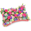 Seen from a slightly elevated, oblique angle against a clean white backdrop, this pillow-shaped floral tribute is a study in colour, texture and quiet dignity. The top surface is densely layered with hot pink roses-velvety, often in bud or just beginning to unfurl-interwoven with soft blush-pink carnations whose ruffled edges create a gentle, tactile surface. Deep purple lisianthus punctuates the composition like small, bell-shaped shadows, while sunny lemon-yellow ranunculus offer bright, buttercup-like accents that lift the palette and suggest warmth. Cool silver-blue Eryngium thistles push through the softness with architectural spikes, their metallic tones echoed by rounded, silvery eucalyptus leaves and a variety of deeper green foliage that give depth and structure beneath the blooms. The cushion rests on a rich, ruffled red satin base, the pleated ribbon framing the rectangle and defining its classic memorial silhouette. Under soft studio light the petals catch a diffuse glow, the roses emitting a subtle fragrance that mingles with the aromatic eucalyptus to produce a calming, restorative scent. Meticulous wiring and foam support lie unseen beneath, providing the gentle, even surface that allows each bloom to breathe and read as one composed expression. Traditionally chosen as a sympathy pillow or memorial cushion, this arrangement leans toward celebration of life rather than sombre mourning-its lively fuchsia, violet, lemon and blush tones convey affection, remembrance and comfort. Designed for placement at a funeral service, graveside or memorial corner, it reflects the careful hand of a local florist who understands how flowers can offer solace in Golders Green and the surrounding Finchley Road neighbourhoods.