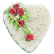 A large heart-shaped sympathy arrangement is set against a crisp white background, its outline clearly defined by a pleated edge of rich dark green satin ribbon. Inside this frame, the heart is fully covered with small, pristine white chrysanthemums, packed closely together to create a smooth, cushion-like surface that looks both soft and carefully structured. The uniform blanket of white blooms gives an impression of purity, stillness, and gentle comfort, like a calm pause amid the sadness of a farewell. Cutting diagonally across the heart is a graceful line of five deep red roses, providing a striking contrast to the white base. At the upper left curve, three of these roses form a focal point, their velvety petals tightly layered and glowing with dark crimson tones. They are nestled among glossy green leaves, fine fern-like greenery, and a scattering of dainty white filler flowers, which add texture and lightness around the richer blooms. Further down and slightly right, two more red roses are spaced apart, their positions balanced and thoughtful. Long, slender strands of bright green ornamental grass loop and arc between the roses, visually joining the upper cluster to the lower flowers and sweeping across the cushion of chrysanthemums with a sense of movement. The whole piece feels traditional yet personal, the kind of floral heart often ordered for services and graveside tributes in and around Golders Green. It expresses deep love, remembrance, and a final, tender message of condolence to a much-missed relative or friend.