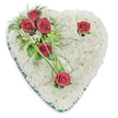 A heart-shaped memorial spray, composed entirely of small, pristine white chrysanthemums, lies against a clean white background like a soft, clouded cushion meant to comfort. Each chrysanthemum bloom is densely set to form a seamless, pillowed surface whose texture reads as quiet and pure; under gentle overhead lighting the petals throw delicate, sighing shadows that give the shape a tender depth. From the upper left a cluster of three deep crimson roses gathers, petals unfurled and velvety, cradled by glossy green leaves and airy, fern-like fronds; fine, arching blades of bright green bear grass and a few tiny white filler flowers gently fan out, adding movement and a natural rhythm across the surface. Two more solitary red roses punctuate the composition-one placed near the right curve of the heart, the other toward the bottom point-each accompanied by modest green accents that let the blooms read as intimate, reflective moments. A narrow emerald-green satin ribbon trims the heart's edge, its sheen defining the perimeter and lending a restrained note of dignity. The scent is implied: the cool, green lift of foliage beneath the soft, powdery chrysanthemum, with the warm, low sweetness of roses threaded through. This is a traditional funeral tribute that speaks of love, respect, and remembrance, crafted with the care of an experienced florist; its calm palette and deliberate placement make it a suitable visual for discussions of sympathy flowers, memorial services, and compassionate floral selections in and around Golders Green and neighbouring Hampstead Garden Suburb.