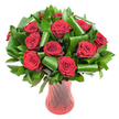 A lush, rounded bouquet of more than a dozen deep crimson roses fills the view, each bloom opened to reveal velvety, tightly spiraled petals that catch the light like soft, satiny folds. The red is rich and almost wine-dark at the petal bases, graduating to a luminous crimson on the outer edges; the even, soft lighting in the photograph brings out the tactile detail of each petal and the subtle sheen that makes the flowers feel freshly cut and tender. Surrounding and supporting the rose heads is an abundant, varied palette of dark green foliage: flat, glossy leaves that form a dense, verdant backdrop, and larger emerald-green leaves artfully rolled and folded into sculptural cylinders and sweeping loops. These shaped green elements - reminiscent of Aspidistra and Ruscus - add volume, modern architecture, and a rhythmic flow that frames each rose like a jewel. The arrangement sits in a tall, slender, translucent ruby-red glass vase with a faint ribbed or twisted texture; the vase's vertical lines catch reflections and echo the passionate red of the blooms while tapering elegantly to a stable base. Set against a pure white background, the bouquet reads as a study in contrast and romance: the saturated reds and deep greens pop with clarity. The overall feeling is intimate and classic, scented as one might imagine with the gentle, heady perfume of fresh roses - ideal for anniversaries, Valentine's, or a refined romantic gesture. This is the kind of centerpiece a local Golders Green florist would recommend for a table at home or a special delivery to a loved one in the neighbourhood.