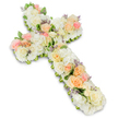 A large, pastel floral cross appears against an immaculate white background, its calm presence evoking the stillness of a chapel in Golders Green before the service begins. The cross is beautifully proportioned and fully clothed in flowers, giving it a soft, almost cushioned appearance. White and creamy ivory blossoms, predominantly roses and generous heads of hydrangea or similar full, rounded blooms, form the base, creating a continuous, cloud-like texture across the vertical stem and horizontal arms. On top of this serene foundation, groups of roses in gentle peach, apricot, and blush pink are positioned at key points along the cross, acting as warm focal accents. Each rose shows delicate, layered petals, some spiralling open in full bloom and others held in a softer, cupped shape, adding subtle variation and life. Throughout the design, small flecks of lavender or lilac filler flowers are woven in, their tiny petals offering light touches of purple that increase the visual depth without disturbing the pastel mood. Fine traces of green foliage peek out along the edges and between certain blooms, underlining the shape of the Christian cross and hinting at renewal and hope. The arrangement is densely constructed, reflecting the practiced hands and respectful approach of a local funeral florist working in the NW11 area. Soft, balanced lighting enhances the gentle colours and casts a faint shadow beneath the cross, giving it a three-dimensional quality on the white ground. The overall impression is one of quiet faith, remembrance, and enduring love, a floral tribute designed to bring comfort and beauty during times of loss.