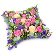 A serene cushion-shaped funeral tribute, composed entirely of closely placed blooms, appears almost like a soft bed of flowers floating against a clean white backdrop. Its form is a neat rectangle with gently curved corners, giving it the comforting silhouette of a pillow laid ready for rest. Across the top, roses in shades of bright fuchsia, tender rose-pink, and creamy pale yellow form the main structure. Some roses are fully open, revealing intricate layers of petals and a velvety texture, while others remain slightly cupped, suggesting a life remembered at different stages. Woven around and between these are velvety purple lisianthus, their deep royal hues adding a sense of depth and quiet dignity to the tribute. Fine clusters of tiny lavender and lilac filler flowers - reminiscent of statice or limonium - thread through every available gap, ensuring the surface is plentiful and full, like a carefully quilted textile. Along the edges and tucked among the blooms, rounded silvery eucalyptus leaves mix with darker, narrow, fern-like greenery, giving the arrangement structure, gentle movement, and a cool, fragrant freshness. The pure white background and subtle shadow beneath the cushion help every colour appear crisp and true, much as it would when arranged by a skilled local florist preparing a tribute for a service in one of Golders Green's chapels or synagogues. The overall impression is one of tenderness and reassurance, a floral pillow designed to convey sympathy, respect, and the wish for peaceful sleep to someone much loved and deeply missed.
