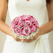 This romantic bridal bouquet, held securely yet tenderly in the bride's hands, is composed of full-headed blush pink roses embraced by airy clouds of white gypsophila. Each rose is at its prime, petals unfurling in layered, silken rings that catch the soft light and reveal faint gradients of pink and cream. The gypsophila, with its countless miniature blooms, fills the spaces between the roses like a delicate mist, smoothing every edge and giving the arrangement an almost ethereal glow. Together they form a neat, rounded posy that feels both classic and modern, the kind of bouquet that suits an elegant ceremony at a Golders Green registry office just as beautifully as a traditional wedding near Hampstead Garden Suburb. The bride's lace gown provides a textured ivory backdrop, its finely worked patterns echoing the tiny blossoms of the baby's breath and drawing the eye to the contrast between fabric and flower. You can almost sense the coolness of the stems wrapped tightly beneath her fingers and the gentle, fresh fragrance rising from the open blooms, a blend of soft rose and crisp greenery. The lighting is natural and flattering, perhaps coming through a synagogue doorway on Russell Parade or a window overlooking Golders Green Road, deepening the shadows between petals and highlighting their velvety sheen. Every element of the bouquet feels carefully judged-balanced, harmonious, and deeply personal-like a floral reflection of the bride's calm joy and quiet anticipation as she prepares to walk down the aisle in this familiar North West London neighbourhood.