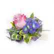A small, exquisitely composed floral corsage or boutonniere lies against a clean white background, its details arranged with the quiet confidence of a seasoned florist. At the heart sits a single rose, partially open, its petals graduating from a soft lavender-pink in the center to a vivid fuchsia at the outer edges, the velvety folds catching a soft, diffused light that suggests early-morning preparation for a wedding or prom. To the right, a compact cluster of indigo-purple florets - reminiscent of tiny hydrangea blooms - offers jewel-like contrast, each petal centring on a bright blue and lime-yellow eye that seems to glimmer against the deeper tones. Surrounding foliage is invitingly varied: broad variegated ivy leaves with deep emerald centres and sunny yellow margins create a classic frame, while slender, silvery-green leaves - like lavender foliage - soften the composition with a muted, tactile hush. A sculptural, striped green-and-cream blade curls elegantly through the bouquet, and wispy asparagus-fern-like sprigs breathe airiness into the whole. The arrangement sits atop a layer of dark purple, web-like mesh that peeks out to add modern structure and depth, catching the light for subtle shimmer. At the lower right a short strand of luminous white pearls suggests a wristlet or decorative pin, lending a touch of tradition. This compact piece radiates romance and craftsmanship, ideal for fastening to a lapel or worn as a wrist corsage at a Golders Hill Park wedding, a school prom, or an intimate celebration locally in Golders Green, where fresh, thoughtful floristry is part of the neighbourhood character.