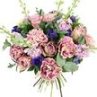 A lavish bouquet is presented against a clean white backdrop, giving the impression of calm gallery light highlighting each flower, much like a still moment in a Golders Green townhouse. At its core, fully opened garden roses in blush pink, dusty mauve, and antique rose tones spiral outward, their velvety petals showcasing a classic romantic elegance. Around them, generous frills of carnations in creamy blush deepen to rose and fuchsia along the ruffled tips, building a padded, almost cloud-like texture that feels soft even to the imagination. Threaded through this gentle palette, intense indigo and royal blue blooms appear as compact, tulip-shaped buds and more open faces, scattering cool, gemstone flashes among the warmer shades. Near the front, a striking royal blue anemone with a dark, nearly black centre anchors the arrangement, drawing the eye like a focal jewel. Slim stems of lilac and pale lavender stock slip upward, their numerous tiny blossoms adding a sense of vertical lift and echoing the delicate, powdery fragrance often noticed in gardens near Golders Hill Park. The whole bouquet is wrapped in fresh greenery: glossy dark green leaves provide structure, while silvery-green eucalyptus, with its rounded leaves and soft tone, blurs the outer edges and hints at a clean, aromatic freshness. The visibly gathered stems at the base reveal its hand-tied nature, suggesting it has just been finished for local delivery in and around Golders Green, Childs Hill, and Cricklewood. With its thoughtful mix of colour and texture, this design feels perfect for anniversaries, heartfelt birthdays, or sending gentle comfort when words are hard to find.