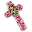 The image features a lush floral cross, meticulously made for a funeral or memorial setting, captured against a stark white background that highlights its gentle pastel tones. The cross itself is fully covered in flowers so that the underlying form is defined only by the arrangement of blooms. At its centre, three prominent roses in soft dusty-rose and muted mauve shades are in full bloom, each petal layered gracefully to create a calm, rounded focal point. Surrounding these roses is a ring of fresh greenery-fine fern fronds and compact green leaves-that introduces contrast and enhances the central cluster. From this point, the arms and stem are richly dressed with lisianthus and other trumpet-shaped blossoms in a variety of pink and lilac-mauve tones, some with slightly darker centres that add visual depth. Scattered throughout are clusters of delicate pink and purple filler flowers, minute white blossoms, and pale green buds, so that no space is left bare, and the overall texture feels plush and continuous. Along the edges, especially toward the tips of the cross, faint glimpses of deeper purple ribbon or structural backing peek through, adding a discreet outline that helps the cross stand out. The effect is serene and reverent, the kind of floral cross families in Golders Green might choose for a church, temple, or graveside tribute, offering a soft yet striking symbol of faith, love, and lasting remembrance during a time of farewell.
